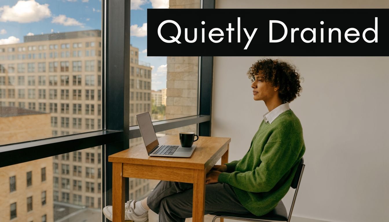A person wearing a green sweater sits at a wooden desk by a window, looking thoughtful and drained.